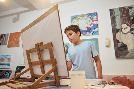 Handsome Caucasian teenage boy drawing on canvas, standing by wooden easel at art class, against the...