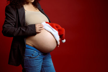 Closeup studio portrait of big belly of a gravid woman, expectant mom putting Santa hat on her pregnant belly, isolated on red studio background with copy ad space. Happy New Year and Merry Christmasの写真素材