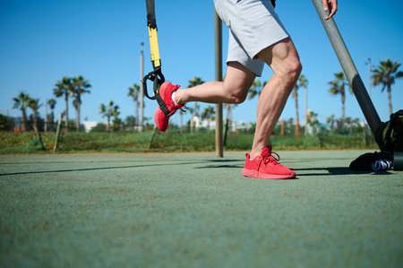 Close-up of a young athletic man in sports clothing training legs with fitness suspension straps in...