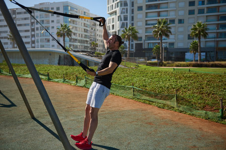 Attractive muscular young adult sportsman, athletic guy exercising with suspension straps outdoors,...