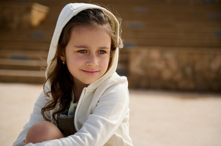 Close-up elementary age Caucasian child, adorable little girl with hood on her head, smiles cutely and dreamily looking away, sitting on skateboard outdoor on urban skatepark against steps backgroundの写真素材