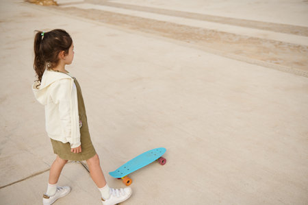Little child girl in beige hoodie and khaki dress, skateboarding on the urban skatepark or playground. View from above. Happy active childhood. Extreme sport. Leisure activityの写真素材