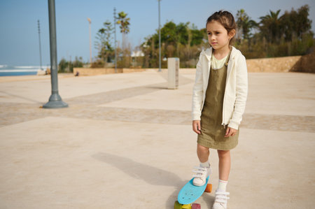 Caucasian cute child girl thoughtfully dreamily looking into the distance, standing on her blue skateboard on one leg on a modern urban skate park. Happy childhood. Healthy active lifestyle conceptの写真素材