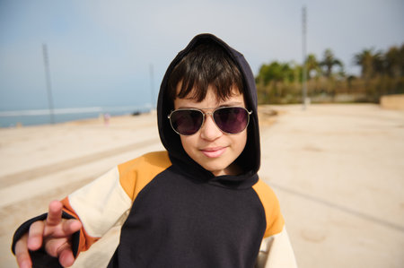 Close-up portrait multi ethnic handsome teenage boy in sunglasses, wearing black hoodie with a hood on his head, smiling looking confidently at camera, riding skateboard in outdoor urban skateparkの写真素材
