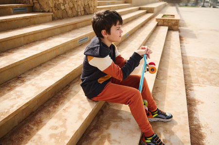 Portrait of a teenage student boy, schoolboy in sportswear sitting on steps in the urban skatepark, thoughtfully looking into the distance. Summer skateboarding, active lifestyle. Sports recreationの写真素材