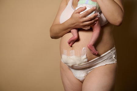 Cropped view of a body positive woman, new mother standing together with her newborn baby. Real body of women after few days of childbirth, isolated on beige studio background. Copy advertising spaceの写真素材