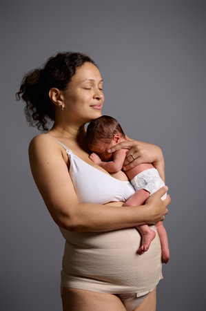 Delightful mixed race woman, happy loving caring mother hugging her newborn baby, standing together isolated gray studio background. Young mom in elastic bandage after cesarean section with her childの写真素材