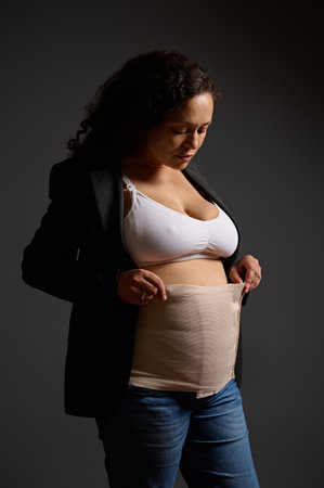 Charming multi ethnic young woman wearing an elastic bandage on her postnatal belly, isolated gray background. Copy space. Postpartum recovery after cesarean c-section. Maternity lifestyle. Body careの写真素材
