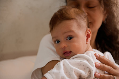 Authentic portrait of adorable baby boy looking at camera, while his mom hugging and holding him in...