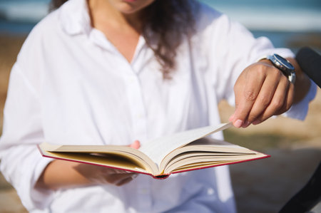 Selective focus on the red hard covered book in the hands of a young woman, leafing through the pages while reading novel on the beach. Digital detox. Literature. People. Lifestyles and hobbiesの写真素材