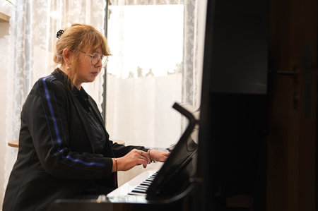 Confident authentic mature Asian woman in black casual attire, playing the piano and singing, teaching piano lesson at homeの写真素材