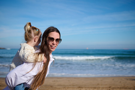 Young woman, happy mother carrying her adorable little kid girl, lovely daughter on her back, giving her a piggyback ride while walking together on the beach. Beautiful waves breaking on the shoreの写真素材