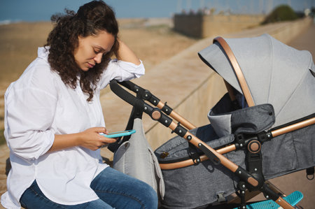Full length young woman, caring mother sharing her maternity lifestyle on social media, sitting on parapet on the beach and pushing a baby pram with cute child sleeping inside outdoor on the beachの写真素材