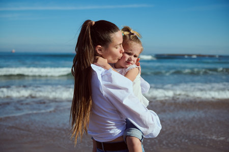 Authentic portrait of a beautiful young Caucasian woman carrying, kissing and hugging her little daughter, enjoying happy time together while walking on the beach. People. Lifestyle. Leisure activityの写真素材