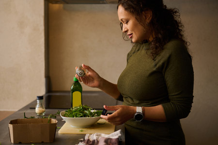 Multi ethnic young adult pretty woman seasoning meal, adding herbs and salt to bowl with salad,...