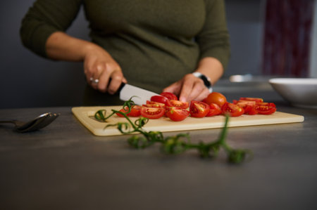Close-up of the hands of a female chef using kitchen knife, chopping tomatoes in two halves on wooden cutting board, standing at kitchen table, preparing healthy delicious meal for lunch or dinnerの写真素材