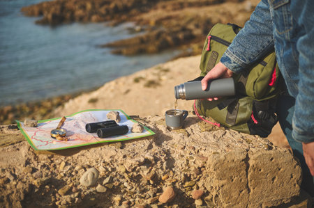 Close-up tourist hand holding flask and pouring hot tea drink into a stainless steel mag, standing on a rock near a map, compass and binoculars. Active lifestyle. Recreation. Tourism conceptの写真素材