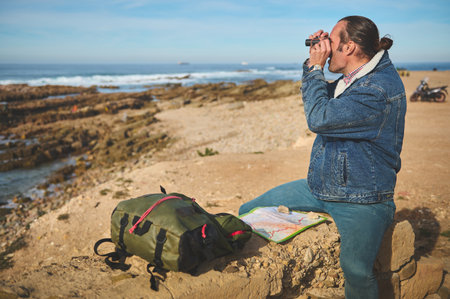 Confident young male adventurer tourist traveler sitting on a rock by sea near his backpack, looking...