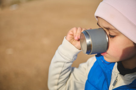 Close-up face portrait of a Caucasian adorable little child girl in pink hat and blue down jacket, drinking hot tea from a steel mug, standing on the nature background. Copy advertising space.の写真素材