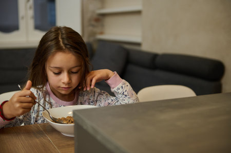 Adorable Caucasian little child girl in pajamas, taking her healthy breakfast at home, eating wholesome flakes with vegetarian milk. Healthy eating. Food and drink consumerism. People. Lifestyleの写真素材