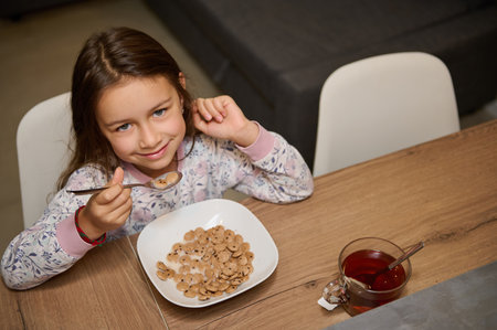 View from above Caucasian adorable little child girl cutely smiling looking at camera, sitting at table and taking her delicious healthy breakfast. Cute kid eating flakes cereals in the home kitchenの写真素材