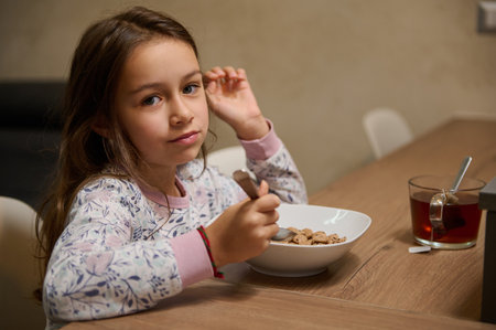 Authentic Caucasian little child girl enjoys eating cereal for morning breakfast with appetite, smiling looking at the camera, sitting at table with a spoon full of oat flakes with milk in her handの写真素材