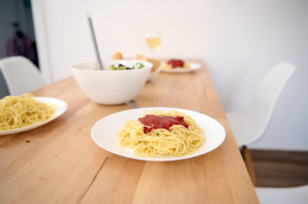 A wooden table with served dishes of freshly prepared Spaghetti with tomato sauce in modern light home kitchen interior. Food and drink concept. Healthy eating and dieting. Italian traditional cuisineの写真素材