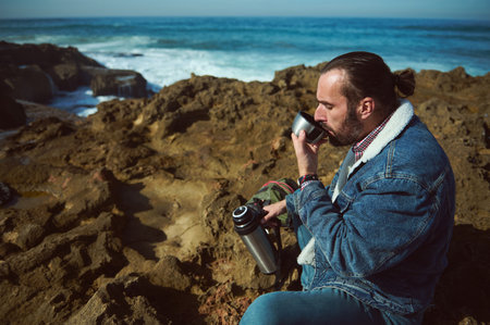 Bearded young man, tourist traveler adventurer sits on the rocky cliff and drinks water from in...