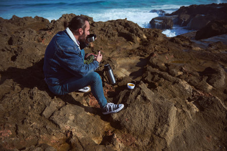 Young brutal bearded man traveling in the wild nature with flask, autumn or spring style, traveler in casual wear have rest, sitting on the rocky cliff by sea. Adventure. Active tourism. Free spiritの写真素材