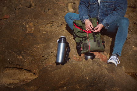 Close-up Caucasian bearded brutal male tourist traveler adventurer with backpack and flask, sitting on the cliff by sea, enjoying his coffee break on the nature. Tourism. Active lifestyle.の写真素材