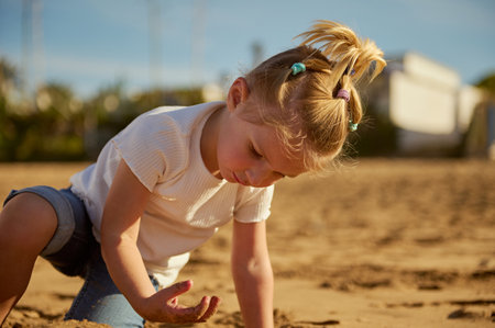 Adorable Caucasian blonde little kid girl playing with wet sand on the beach, standing barefoot and leaving footsteps on the wet sandy beach. People. Active healthy lifestyle. Happy carefree childhoodの写真素材