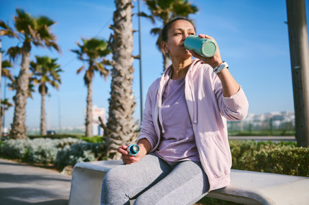 Charming young female athlete drinks water from a sports bottle, sitting on a stone bench against...