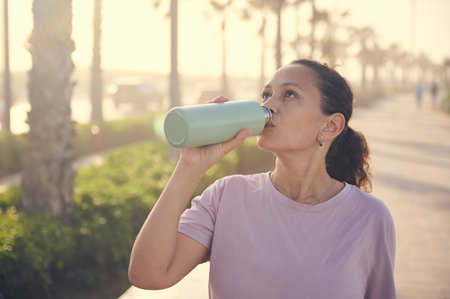 Curly haired Latina woman in purple t-shirt, drinking water from a steel bottle, standing on the...