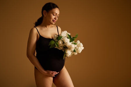 Attractive young adult woman, confident and happy pregnant mother, gravid female in black bodysuit, isolated over beige studio background. Pregnancy. Childbearing. Maternity and parenting conceptの写真素材