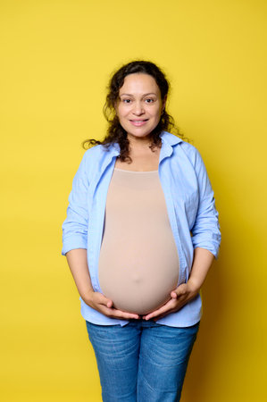 Authentic emotional portrait of a mid adult pregnant woman holding her big belly, smiling looking at camera, isolated yellow background. Pregnancy. Maternity leave. Women's fertility and healthの写真素材
