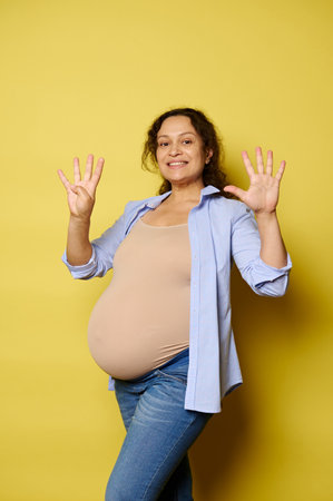 Smiling pregnant woman expecting a baby, showing nine fingers at the camera, isolated on yellow studio background. She's in last trimester of her happy pregnancy. Childbearing. Childbirth. Maternity.の写真素材