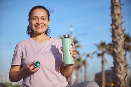 Athletic woman in purple t-shirt holding a bottle of water and smiling looking confidently at...