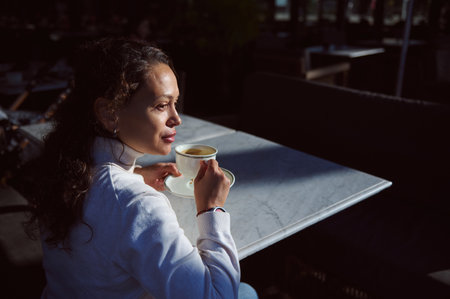 Confident and attractive curly haired young woman looking aside while sitting at coffee shop, enjoying her coffee break. People. Food and drink consumerism conceptの写真素材