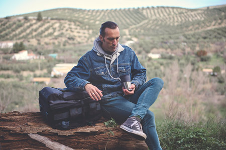 Backpacked adventurer traveler man sitting on a log in mountains, drinking his morning coffee from cup, relaxing on the nature. People. Healthy lifestyle. Weekend leisure activityの写真素材