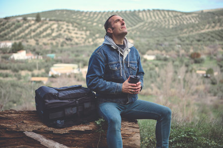 Happy young adult traveler adventurer man with backpack, sitting on a log in the forest, drinking coffee from a stainless steel mug, dreamily looking up, breathing fresh mountains airの写真素材