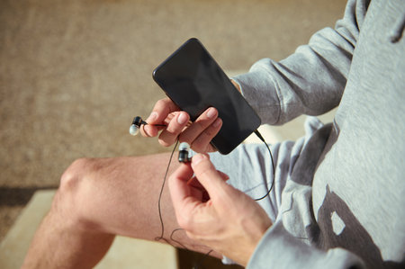 Close-up male athlete hands holding earphones and smartphone with black blank mockup screen. Copy...