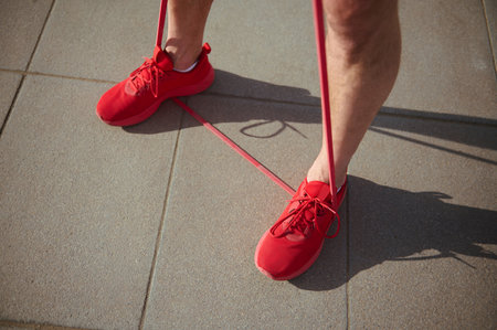 View from above athletic man in red sports shoes, doing physical exercises with red resistance band....