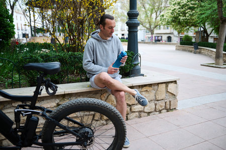 Caucasian young man cyclist in sportswear, drinking water from sports bottle in the city, sitting on a bench, relaxing after riding an eco-friendly e-bike motorbike. Electric bicycle in the foregroundの写真素材