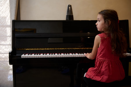 Confident portrait of an inspired beautiful talented little child girl learning music, performing the rhythm of classical music while playing piano, putting fingers on keys and dreamily looking awayの写真素材