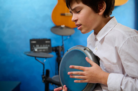 Close-up portrait of teen boy with eyes closed, playing the djembe drum in a cozy home music studio,...