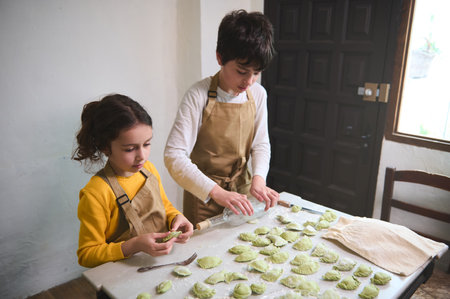 Cute children in the rural kitchen, rolling out dough with a wine bottle, sculpting dumplings with...