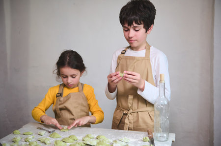 Cooking class for kids. Two diverse kids, a boy and a girl preparing family dinner, standing at floured kitchen table and modeling dumplings or Ukrainian varennyky in the rural house kitchen interiorの写真素材