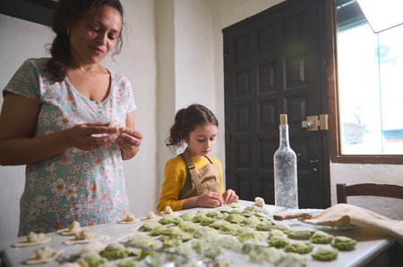 Authentic beautiful young woman, mother and her little kid, daughter in the rural kitchen, sculpting dumplings from dough with mashed potatoes filling. Cooking delicious homemade vegetarian dumplingsの写真素材