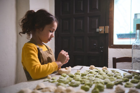 Confident portrait of cute little girl in beige apron, standing at kitchen table with molded dumplings, stuffing the dough and molding ravioli, helping her mother in culinary, preparing dinner aloneの写真素材