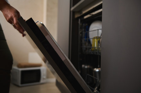 Close-up housewife's hand opening or closing the door of a modern automatic dishwasher, unloading, emptying out, taking out clean dishes, standing in stylish minimalist gray color kitchen interiorの写真素材